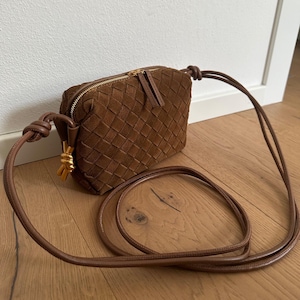 May include: A brown suede crossbody bag with a woven design. The bag has a gold zipper and a long, thin strap. The strap is knotted at the ends. A small gold tassel hangs from the side of the bag. The bag is sitting on a wooden floor.