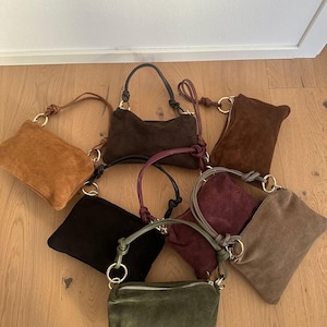 Pode incluir: Uma coleção de bolsas de camurça em várias cores, incluindo castanho, preto, verde-oliva e bordô. Cada bolsa apresenta uma alça superior ou alça e um detalhe de anel de metal. As bolsas estão dispostas sobre uma superfície de madeira.