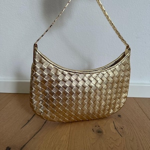 May include: A gold-coloured, woven-style handbag with a curved shape and a matching gold chain strap. The bag is held up by a hand, showcasing its metallic sheen and textured surface. The bag's design features a repeating woven pattern.