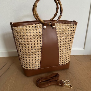 May include: A woven rattan tote bag with brown leather detailing and a bamboo handle. It features a detachable brown leather shoulder strap with gold-coloured clasps. The bag is on a wooden floor.
