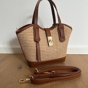 May include: A structured handbag featuring a woven tan straw body with brown leather trim, two top handles, and a gold-tone metal turn-lock closure. The bag includes a detachable brown leather shoulder strap with gold-tone metal lobster clasps.
