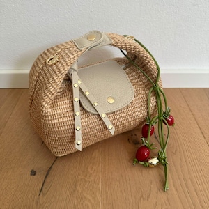 Könnte beinhalten: Eine Handtasche aus geflochtenem Stroh mit einer beigefarbenen Lederklappe und einem Griff. Die Tasche hat goldfarbene Beschläge und einen dekorativen Kirschstiel mit roten Kirschen. Die Tasche steht auf einem Holzboden.