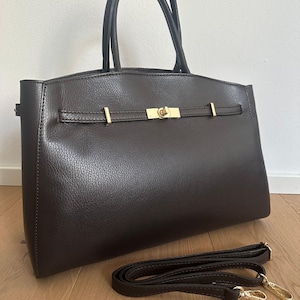 May include: A dark brown leather handbag with a top handle and a detachable shoulder strap. The bag features gold-colored hardware, including a clasp closure and strap attachments. The bag is sitting on a wooden floor.