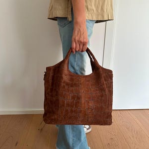 May include: A large, rectangular brown suede tote bag featuring a textured, embossed crocodile skin pattern. The bag has two sturdy top handles and a spacious main compartment, showcasing a rich, matte finish with visible stitching details.