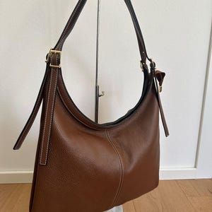 May include: A brown leather shoulder bag with a curved design. The bag features a long, adjustable strap and gold-colored hardware. White stitching accents the seams. The bag is displayed on a metal stand against a white background.