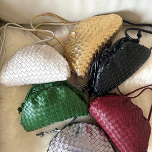 May include: A collection of woven leather clutch bags in various colors, including white, gold, black, green, burgundy, and silver. Each bag features a drawstring closure and a thin shoulder strap. The bags are arranged on a white fur-like surface.