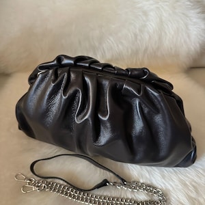 Puede incluir: Un clutch de cuero negro con un diseño fruncido. El bolso tiene una correa de cadena plateada con cierres de mosquetón. El clutch está sobre una superficie blanca y esponjosa.