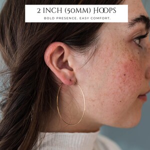 Puede incluir: Un pendiente de aro dorado fino que mide 50 mm de di&aacute;metro. El texto superpuesto dice 2 INCH (50MM) HOOPS y BOLD PRESENCE. EASY COMFORT. El pendiente tiene un dise&ntilde;o circular simple de alambre met&aacute;lico con un acabado pulido.