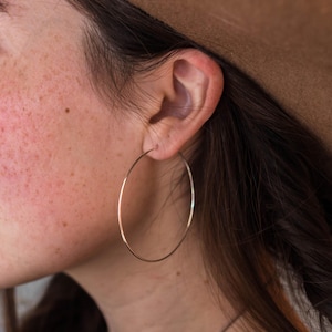 May include: A large, thin, circular hoop earring made of polished gold-toned metal. The earring features a simple, continuous wire design that forms a wide, open circle, hanging from the earlobe with a smooth, reflective surface finish.