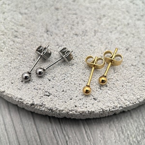 May include: Two pairs of stud earrings. One pair is silver-colored, and the other is gold-colored. Each earring has a small, round ball at the end of a post. The earrings are displayed on a textured, light gray surface.