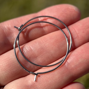 May include: A pair of silver-colored hoop earrings. The earrings are circular, with a simple, minimalist design. The hoops are held in a hand, with a blurred green background.