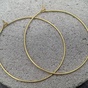 Könnte beinhalten: Ein Paar goldfarbene Creolen. Die Ohrringe sind dünn und kreisförmig, mit einem schlichten, eleganten Design. Die Reifen werden vor einem strukturierten, grauen Hintergrund gezeigt.