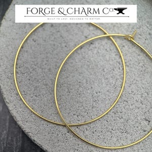 Puede incluir: Dos aros dorados delgados con un dise&ntilde;o de alambre circular. El texto FORGE & CHARM CO. BUILT TO LAST. DESIGNED TO MATTER. es visible en la parte superior de la imagen, acompa&ntilde;ado de un peque&ntilde;o icono de yunque. Los pendientes tienen un cierre simple.