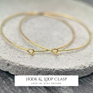 Puede incluir: Dos brazaletes finos de alambre en tono dorado con un mecanismo de cierre de gancho y bucle. Las pulseras circulares est&aacute;n construidas con un material de alambre met&aacute;lico liso. El texto Hook & Loop Clasp Lock In. Stay Secure. aparece debajo.