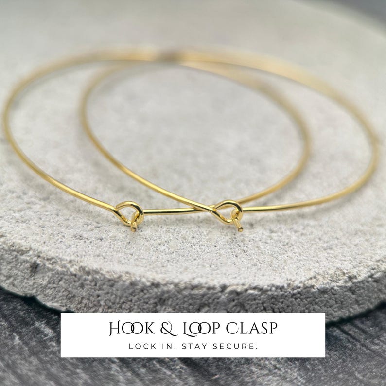 Puede incluir: Dos brazaletes finos de alambre en tono dorado con un mecanismo de cierre de gancho y bucle. Las pulseras circulares est&aacute;n construidas con un material de alambre met&aacute;lico liso. El texto Hook & Loop Clasp Lock In. Stay Secure. aparece debajo.
