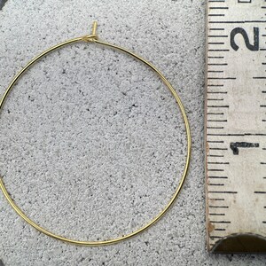 Puede incluir: Un pendiente de aro circular fino de tono dorado con un peque&ntilde;o cierre de bucle en la parte superior. El aro est&aacute; colocado junto a una regla de madera, mostrando un di&aacute;metro de aproximadamente 3,8 cent&iacute;metros desde el borde exterior.
