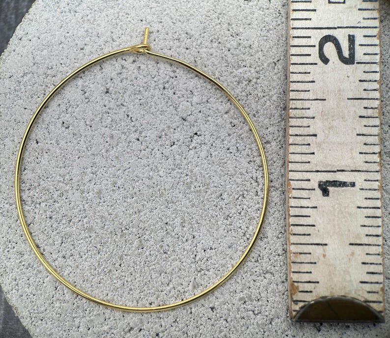 Puede incluir: Un pendiente de aro circular fino de tono dorado con un peque&ntilde;o cierre de bucle en la parte superior. El aro est&aacute; colocado junto a una regla de madera, mostrando un di&aacute;metro de aproximadamente 3,8 cent&iacute;metros desde el borde exterior.