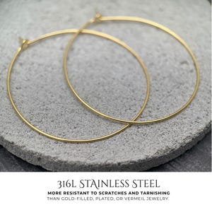Puede incluir: Un par de aros dorados finos hechos de acero inoxidable 316L. El texto debajo de los pendientes dice: 316L STAINLESS STEEL. MORE RESISTANT TO SCRATCHES AND TARNISHING THAN GOLD-FILLED, PLATED, OR VERMEIL JEWELRY.