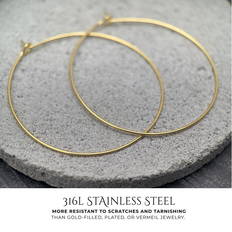 Puede incluir: Un par de aros dorados finos hechos de acero inoxidable 316L. El texto debajo de los pendientes dice: 316L STAINLESS STEEL. MORE RESISTANT TO SCRATCHES AND TARNISHING THAN GOLD-FILLED, PLATED, OR VERMEIL JEWELRY.
