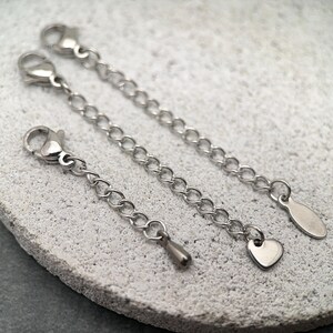 May include: Three silver-colored chain extenders with lobster claw clasps. One extender has a heart-shaped charm, another has a teardrop charm, and the third has a simple oval charm. The extenders are on a textured, light gray surface.