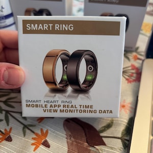 Pode incluir: Uma caixa com a etiqueta "SMART RING" mostra dois anéis inteligentes, um dourado e outro preto. O texto na caixa diz "SMART HEART RING, MOBILE APP REAL TIME, VIEW MONITORING DATA". Outras caixas são visíveis no fundo.