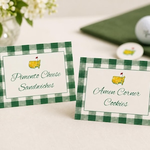 May include: Two place cards with green and white gingham borders. The cards display the text "Pimento Cheese Sandwiches" and "Amen Corner Cookies" with a golf course logo. A golf ball and a green napkin are in the background.