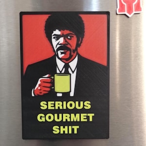 May include: A black rectangular magnet featuring a stylized image of a man in a suit holding a yellow mug. The text "SERIOUS GOURMET SHIT" is in yellow block letters below the image.