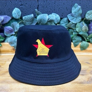 May include: A black bucket hat with a red star and a yellow bird emblem. The hat has a classic bucket shape and is made of a dark fabric. The hat is a stylish accessory, suitable for various outfits.