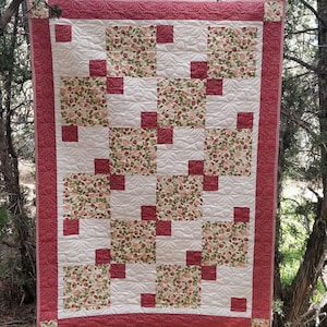 Pode incluir: Um edredom feito à mão com um design de patchwork. O edredom apresenta quadrados alternados de tecido branco, estampa floral e quadrados vermelhos sólidos. A borda é vermelha sólida. O edredom está pendurado ao ar livre, mostrando suas cores e texturas.