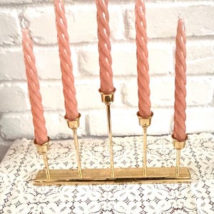 May include: A brass candle holder with five pink twisted candles. The candles are arranged in a row, each held by a slender gold stem. The holder sits on a table covered with a white lace tablecloth, against a white brick wall.