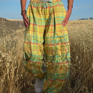 May include: Yellow, orange, and teal plaid pants with a drawstring waist. The pants have a loose, baggy fit and are worn with white sneakers. The image is taken outdoors in a field of tall grass.