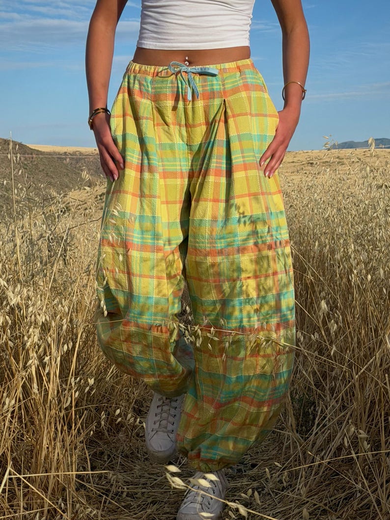 May include: Yellow, orange, and teal plaid pants with a drawstring waist. The pants have a loose, baggy fit and are worn with white sneakers. The image is taken outdoors in a field of tall grass.