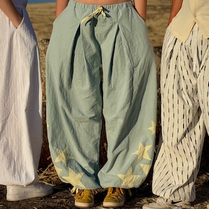 May include: Light blue pants with yellow star appliques. The pants have a wide, loose fit and a drawstring waist. The image also shows a white dress and striped pants.