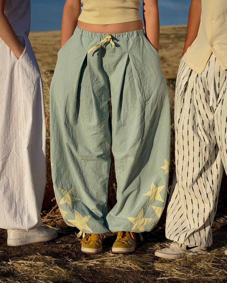 May include: Light blue pants with yellow star appliques. The pants have a wide, loose fit and a drawstring waist. The image also shows a white dress and striped pants.