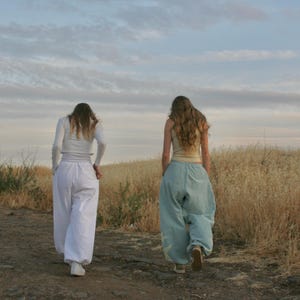 May include: Two people walking away from the camera on a dirt path. One wears a white long-sleeved top and white pants. The other wears a light-colored tank top and light blue pants. The background features dry grass and a cloudy sky.