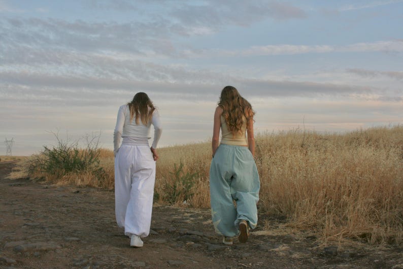 May include: Two people walking away from the camera on a dirt path. One wears a white long-sleeved top and white pants. The other wears a light-colored tank top and light blue pants. The background features dry grass and a cloudy sky.