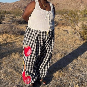 May include: A white crochet tank top with a square neckline and a drawstring closure is paired with black and white checkered pants. The pants feature two large red strawberry appliqu&eacute;s. The person is wearing black sandals.