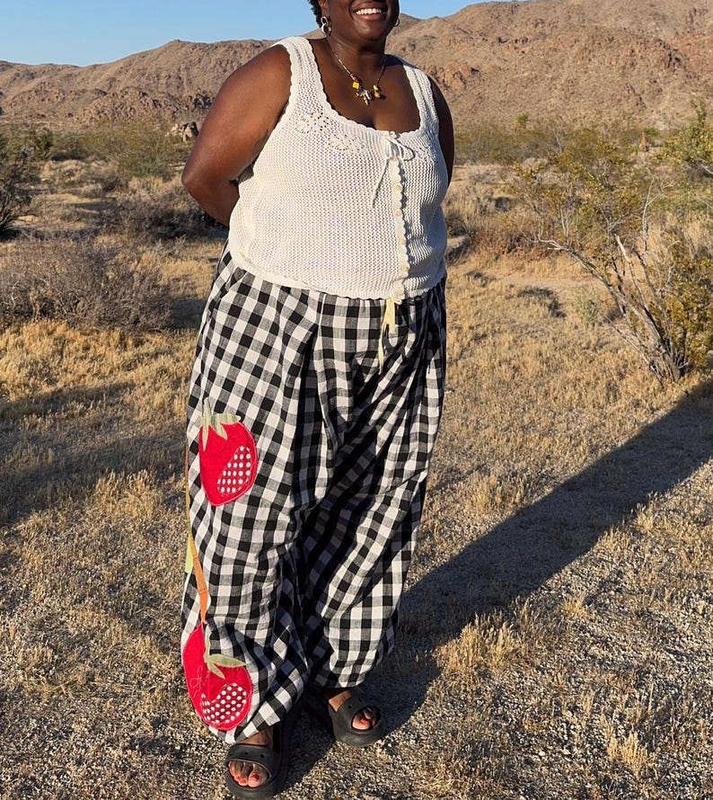 May include: A white crochet tank top with a square neckline and a drawstring closure is paired with black and white checkered pants. The pants feature two large red strawberry appliqu&eacute;s. The person is wearing black sandals.