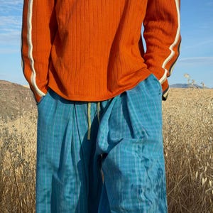 May include: An orange ribbed long-sleeve shirt with white and black stripes on the sleeves. The person is wearing blue plaid pants with a yellow drawstring. The outfit is set against a backdrop of dry grass and a blue sky.