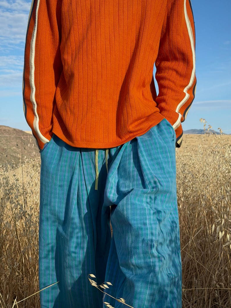 May include: An orange ribbed long-sleeve shirt with white and black stripes on the sleeves. The person is wearing blue plaid pants with a yellow drawstring. The outfit is set against a backdrop of dry grass and a blue sky.