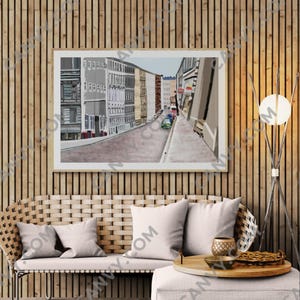 Könnte beinhalten: Ein gerahmter Kunstdruck, der eine Straßenszene mit Gebäuden und Autos zeigt. Das Kunstwerk wird über einem Korbsessel mit weißen Kissen und einem passenden Hocker ausgestellt. Ein Holztablett mit Dekoration steht auf dem Hocker, und rechts befindet sich eine moderne Stehlampe.