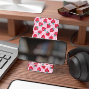 Può includere: Un supporto per telefono con un motivo a fragole rosa. Il supporto contiene uno smartphone nero e il design presenta fragole rosse e fiori bianchi su sfondo rosa. Il supporto è realizzato in un materiale robusto ed è progettato per tenere un telefono in posizione verticale.