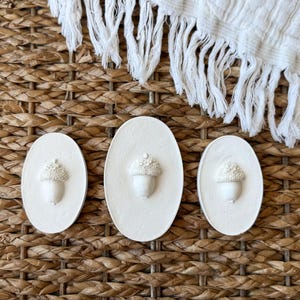 May include: Three white, oval-shaped decorative pieces with raised acorn designs. The acorns have textured caps and smooth bodies. The pieces are arranged on a woven, brown surface, with a white fringed cloth in the background.