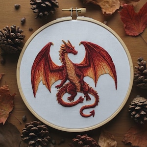 Puede incluir: Un bastidor de bordado circular de madera que contiene tela blanca con un diseño detallado cosido con hilo de un dragón rojo y naranja. El dragón se muestra con grandes alas extendidas, una cola larga y escamas texturizadas en tonos carmesí.