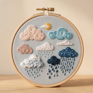 May include: A circular wooden embroidery hoop containing light grey fabric with hand-stitched clouds and rain. The design features pink, blue, white, and dark navy clouds, a yellow sun, and vertical blue stitches representing falling rain.