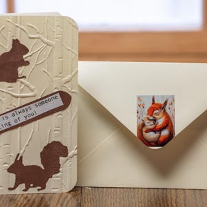 May include: A greeting card with a textured, beige background featuring embossed squirrels and birch tree designs. The card includes a brown label with the text "There is always someone thinking of you!". An envelope with a squirrel sticker is next to the card.