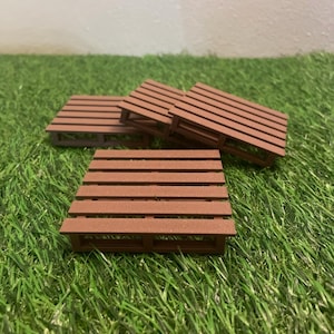 Peut inclure: Quatre mini-palettes en bois marron disposées sur un lit d'herbe verte artificielle. Les palettes sont rectangulaires avec des lattes horizontales et une structure de support simple. Les palettes sont de couleur marron chaud.