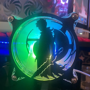 Puede incluir: Un ventilador de ordenador negro con un diseño de silueta de un personaje sosteniendo una espada. El ventilador tiene un centro transparente con iluminación LED verde y azul. Fijado con tornillos plateados.