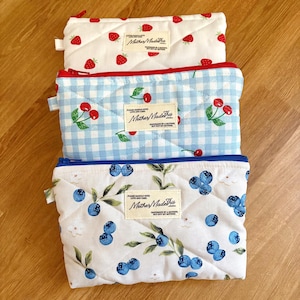 May include: Three rectangular quilted zipper pouches featuring fruit prints. The top pouch is white with red strawberries, the middle is light blue gingham with red cherries, and the bottom is white with blue blueberries. Each has a MotherMadeThis label.