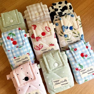 May include: A collection of quilted fabric pouches featuring various patterns including blue and white gingham with cherries, strawberries on cream, blueberries, cow print, and polka dots. Each pouch has a flap with a snap button and a label.